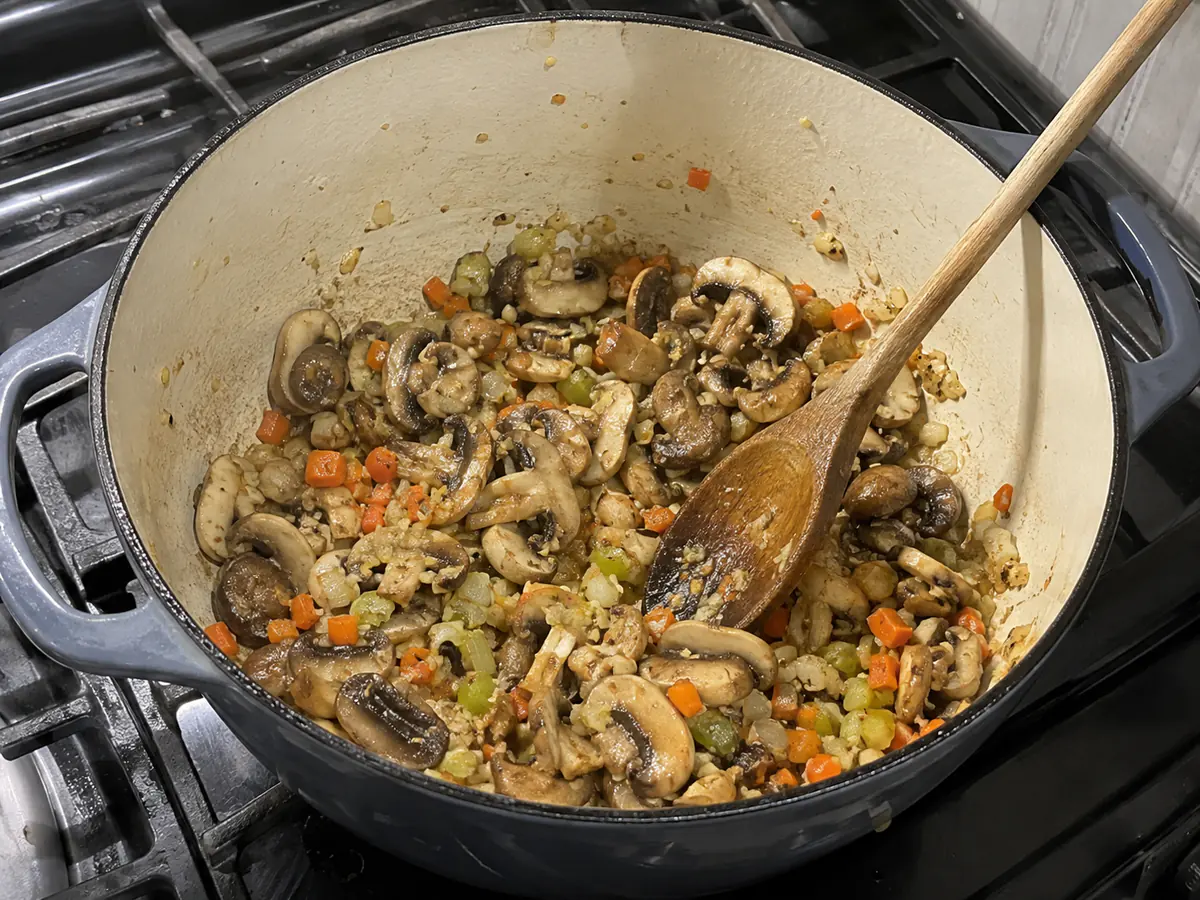 Sliced cremini mushrooms browning with softened onion, carrots, celery, and garlic in a Dutch oven.