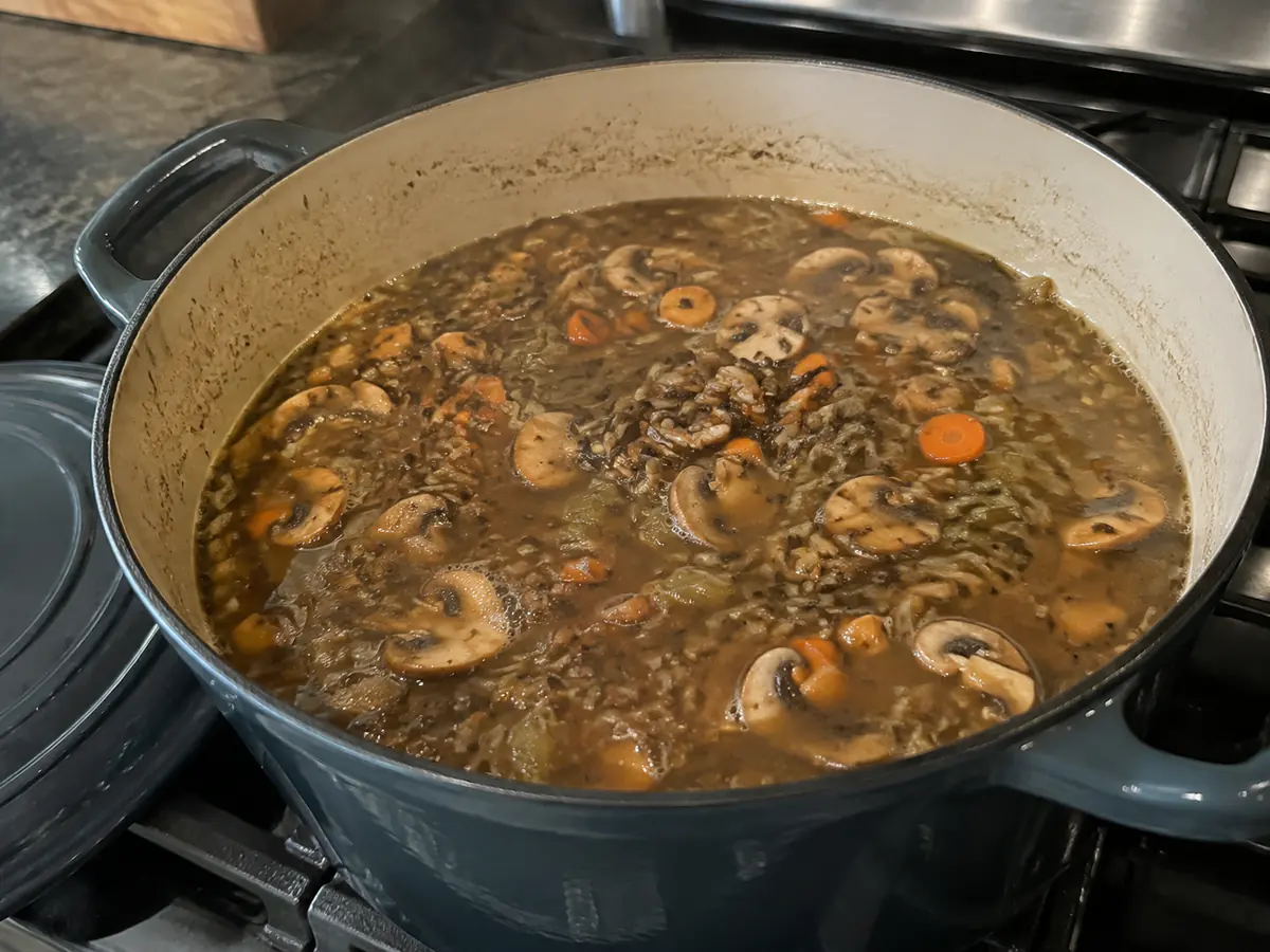 Wild rice, mushrooms, and vegetables simmering in broth in a dark enameled Dutch oven.
