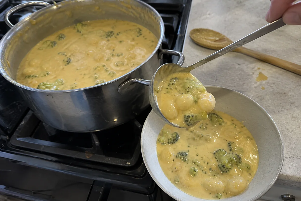 Broccoli cheddar gnocchi soup being ladled from a stainless-steel pot into a ceramic bowl.