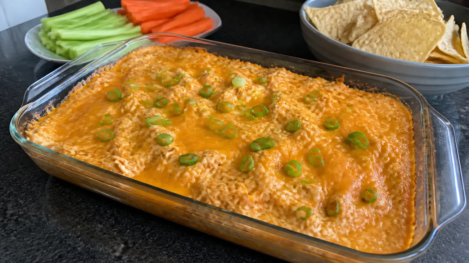 Baked buffalo chicken dip topped with melted cheddar and green onions beside celery, carrots, and tortilla chips