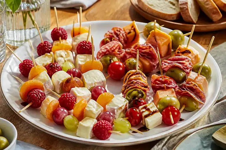 Mixed sweet and savory cheese skewers with fruit, olives, tomatoes, salami, and prosciutto on a party platter.