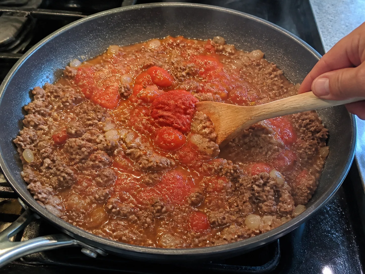 Tomato paste and diced tomatoes being stirred into browned ground beef and onions.