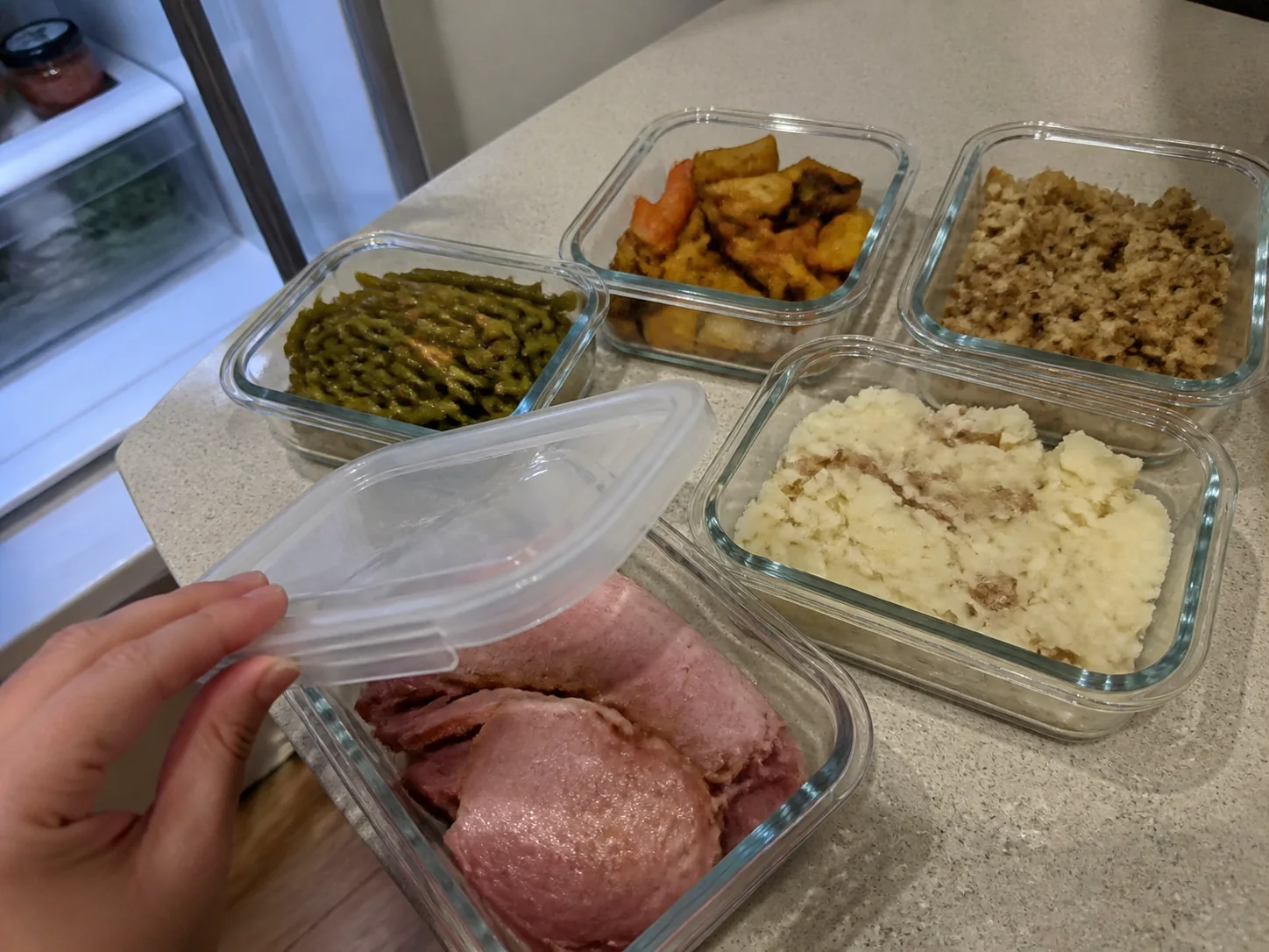 Christmas dinner leftovers portioned into clear containers with ham, potatoes, vegetables, and stuffing.