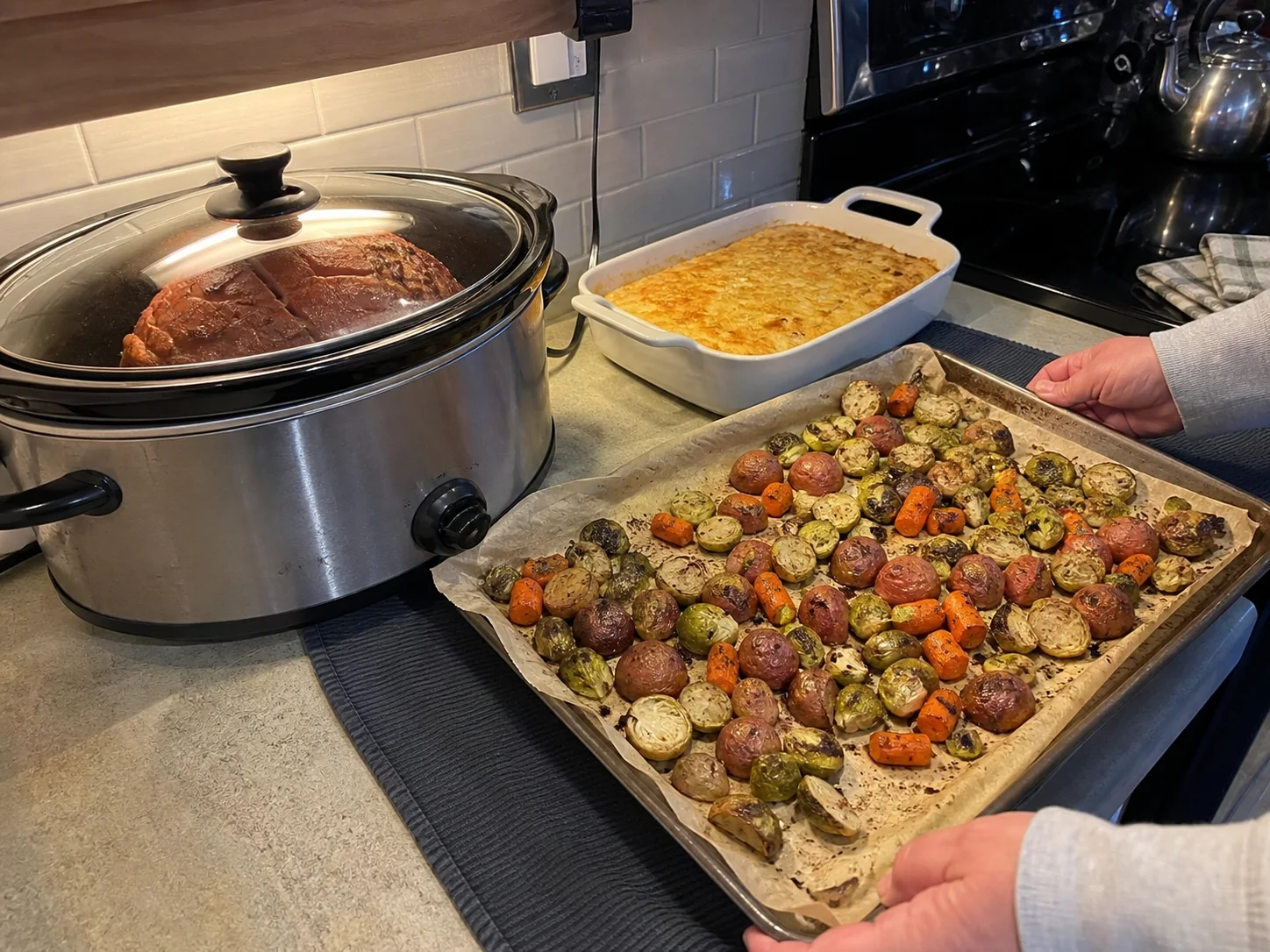 Home cook using a slow cooker for ham while vegetables roast on a sheet pan beside the oven.