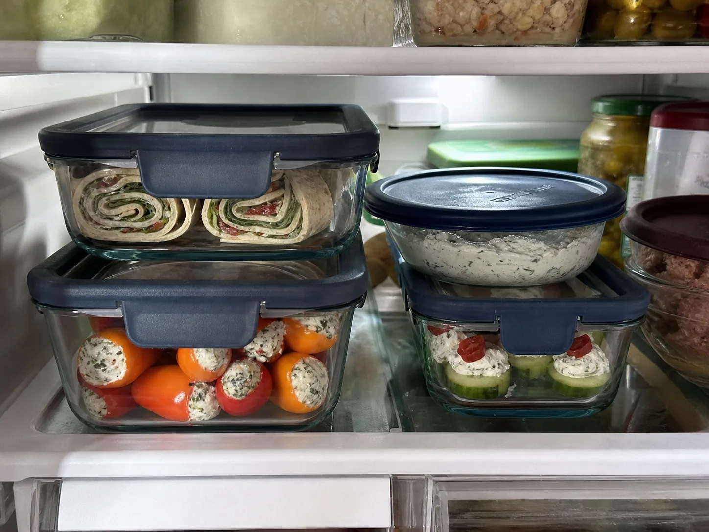 Pinwheels, herb cream cheese dip, stuffed mini peppers, and cucumber bites stored in covered refrigerator containers.