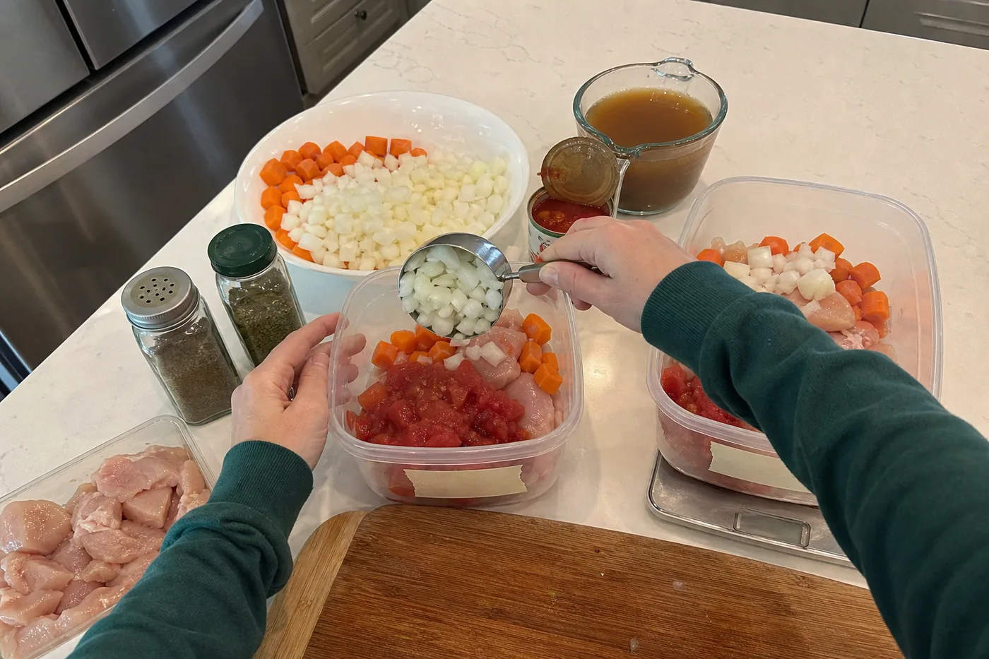 Thawed chicken, carrots, onions, tomatoes, broth, and spices being portioned into meal-prep containers.