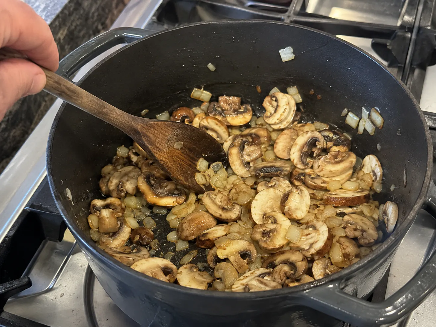 Sliced mushrooms browning with softened onion in a charcoal Dutch oven before broth is added.