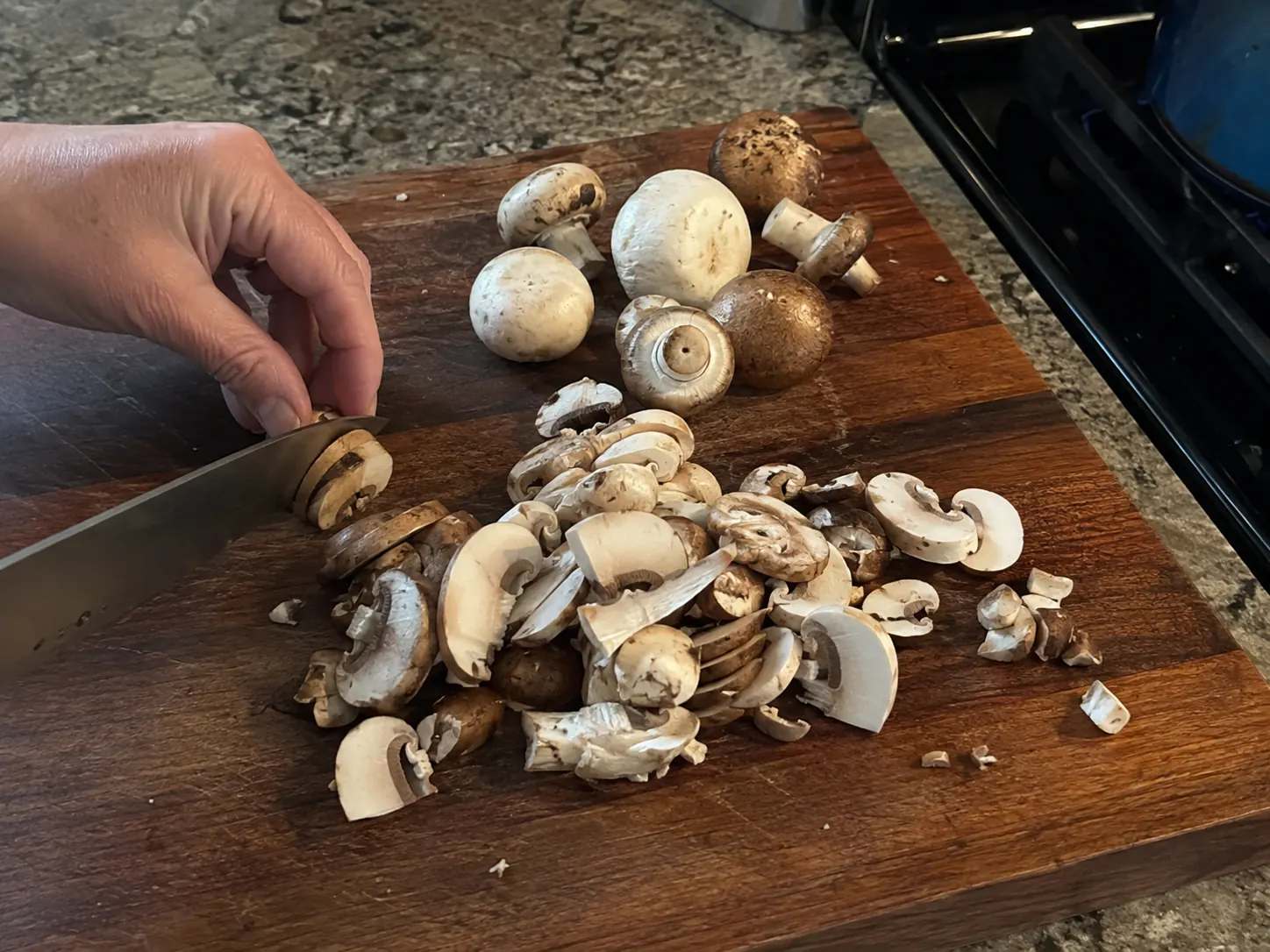 Cremini and white button mushrooms being sliced on a wooden board for homemade mushroom soup.