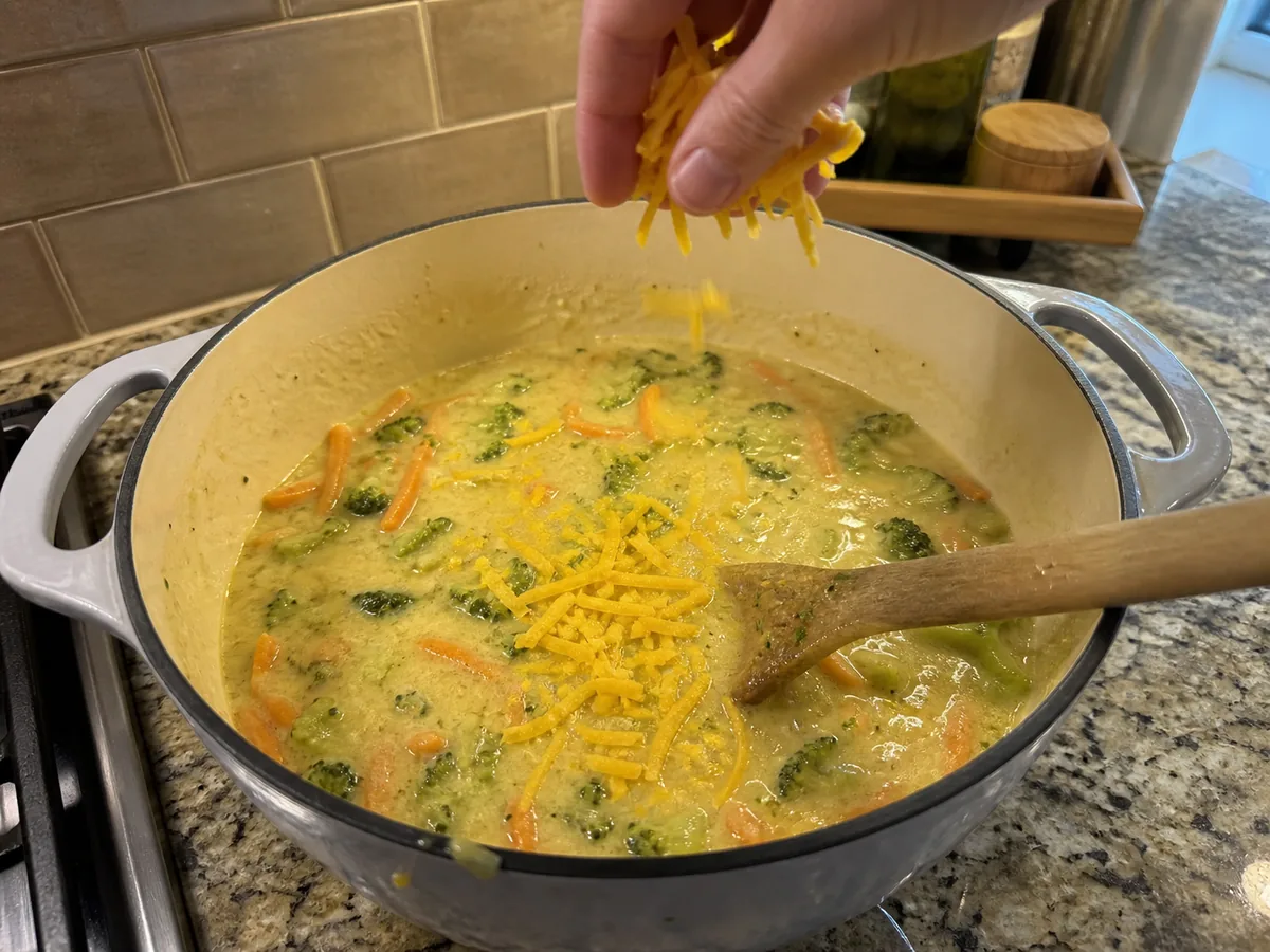 Freshly grated sharp cheddar being stirred into broccoli soup off the heat.